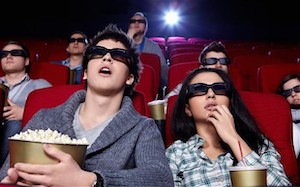 A couple sitting together in a cinema watching a film in a cinema with 3D glasses whilst eating popcorn and drinking soda from gold colored containers whilst others behind them look on ... that said, one looks like they are sleeping ...